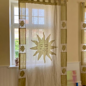 Könnte beinhalten: Cremefarbener Vorhang mit einem großen Sonnendesign in der Mitte. Die Sonne hat ein lächelndes Gesicht. Der Vorhang hat olivgrüne Ränder und sternförmige Akzente. Der Vorhang hängt vor einem Fenster.