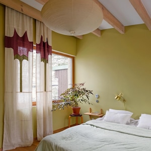 K&ouml;nnte beinhalten: Ein Schlafzimmer mit cremefarbenen Vorh&auml;ngen mit dunkelrotem und gr&uuml;nem geometrischem Muster, einer gro&szlig;en beigen Papier-Pendelleuchte, einer hellgr&uuml;nen Tagesdecke mit wei&szlig;en Kissen, einer Topfpflanze auf einem Hocker und einer Messing-Wandleuchte.