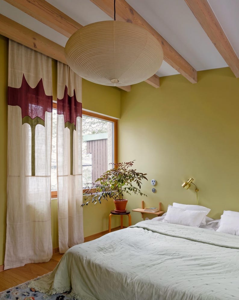 K&ouml;nnte beinhalten: Ein Schlafzimmer mit cremefarbenen Vorh&auml;ngen mit dunkelrotem und gr&uuml;nem geometrischem Muster, einer gro&szlig;en beigen Papier-Pendelleuchte, einer hellgr&uuml;nen Tagesdecke mit wei&szlig;en Kissen, einer Topfpflanze auf einem Hocker und einer Messing-Wandleuchte.