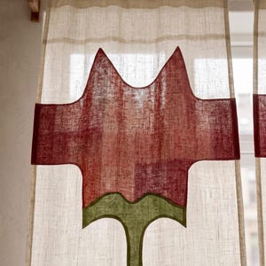 K&ouml;nnte beinhalten: Ein halbtransparenter, cremewei&szlig;er Vorhang an einer Holzstange mit einem gro&szlig;en, mittig applizierten Design einer stilisierten roten Blume mit gr&uuml;nem Stiel und Bl&auml;ttern, gefertigt aus strukturiertem Stoff mit sichtbarem Webmuster.