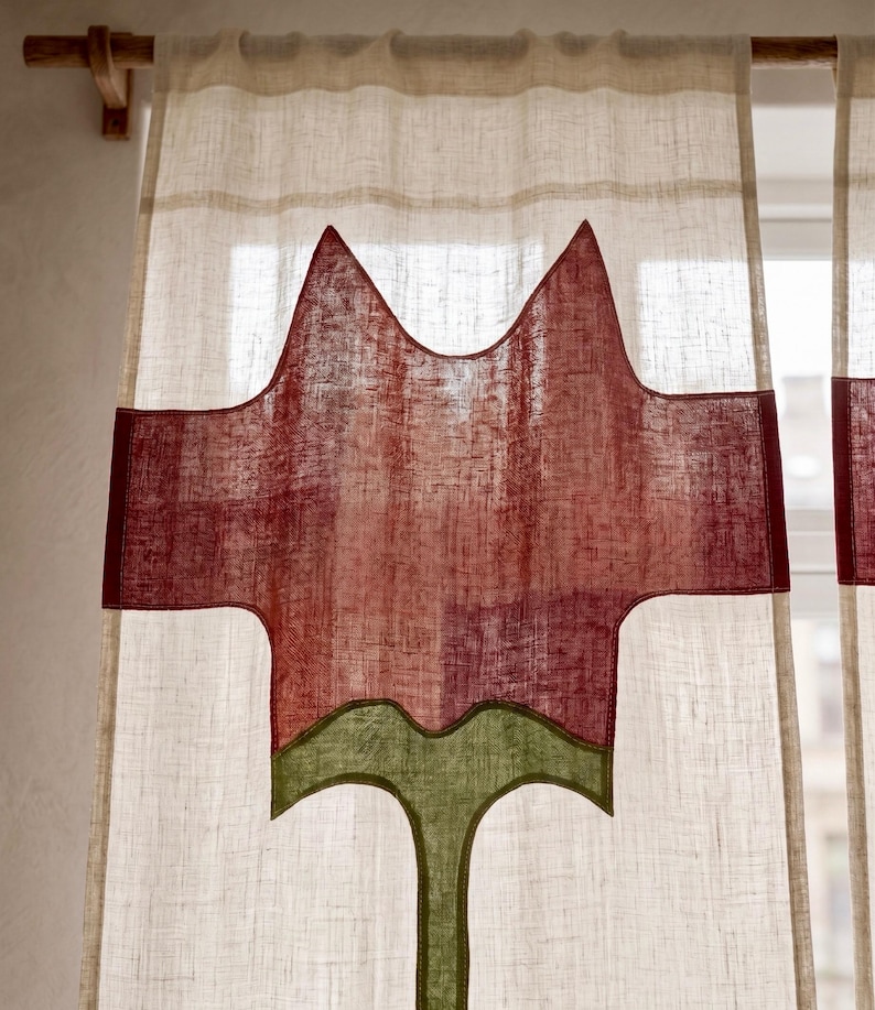 K&ouml;nnte beinhalten: Ein halbtransparenter, cremewei&szlig;er Vorhang an einer Holzstange mit einem gro&szlig;en, mittig applizierten Design einer stilisierten roten Blume mit gr&uuml;nem Stiel und Bl&auml;ttern, gefertigt aus strukturiertem Stoff mit sichtbarem Webmuster.