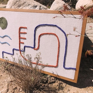 May include: A framed wall art piece with a white canvas background and colorful yarn embroidery. The design features a blue and orange curved line, a blue wavy line, and a green circle.