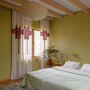 K&ouml;nnte beinhalten: Ein Schlafzimmer mit olivgr&uuml;nen W&auml;nden, einer hellgr&uuml;nen Tagesdecke und wei&szlig;en Vorh&auml;ngen mit einem rot-gr&uuml;nen abstrakten Muster. Eine gro&szlig;e beige Pendelleuchte h&auml;ngt an h&ouml;lzernen Deckenbalken &uuml;ber einem Bett mit wei&szlig;en Kissen und einem gemusterten Teppich.