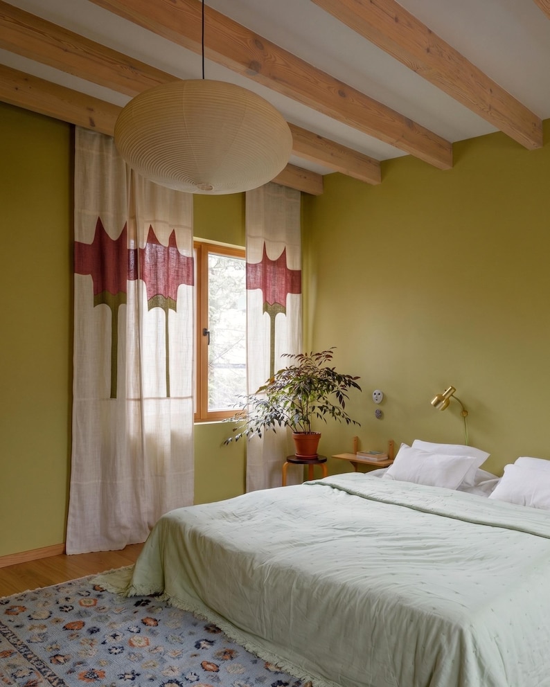 K&ouml;nnte beinhalten: Ein Schlafzimmer mit olivgr&uuml;nen W&auml;nden, einer hellgr&uuml;nen Tagesdecke und wei&szlig;en Vorh&auml;ngen mit einem rot-gr&uuml;nen abstrakten Muster. Eine gro&szlig;e beige Pendelleuchte h&auml;ngt an h&ouml;lzernen Deckenbalken &uuml;ber einem Bett mit wei&szlig;en Kissen und einem gemusterten Teppich.