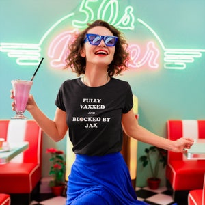 May include: A woman wearing a black t-shirt with the text "FULLY VAXxed AND BLOCKED BY JAX" in white. She is smiling and holding a pink milkshake in a glass with a straw. She is standing in a retro diner setting.