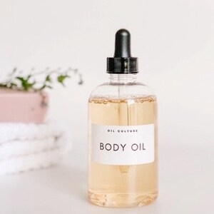 May include: A clear glass bottle with a black dropper top, filled with light amber body oil. The bottle has a white label with the text "OIL CULTURE" above "BODY OIL". A stack of white towels and a pink bar of soap are in the background.