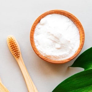 Puede incluir: Un cuenco de madera lleno de polvo blanco está junto a un cepillo de dientes de bambú. Una hoja verde está en la esquina. El cepillo de dientes tiene cerdas de color marrón claro. El cuenco es de madera marrón claro.