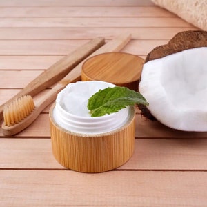 May include: A natural skincare arrangement featuring a jar of cream with a mint leaf, two wooden toothbrushes, a coconut half, and a rolled towel. The cream jar has a bamboo exterior. The items are arranged on a light wooden surface.