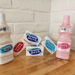 以下が含まれることがあります： ZOTEの洗濯製品の品揃え。白とピンクの液体洗剤と石鹸バーが含まれています。ボトルと石鹸バーには、青と赤のZOTEロゴと「Blanco」と「Rosa」の文字が表示されています。