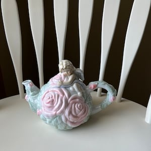 May include: A porcelain teapot shaped like a rose bush with a cherub sitting on top. The teapot is decorated with pink roses and green leaves.