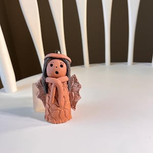 May include: A terracotta-colored angel figurine with a surprised expression. The angel has dark hair, a matching hat, and a scarf. It features textured wings and a patterned dress. The figurine is displayed on a white surface.