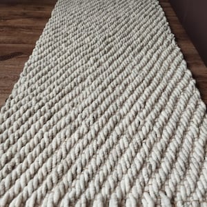 May include: A rectangular, off-white rug with a thick, braided texture. The rug has a diagonal pattern and a fringe along the shorter ends. The rug is placed on a wooden floor.