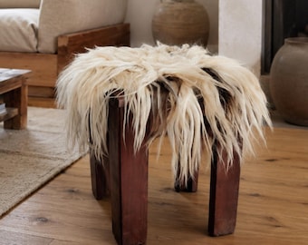 Op de afbeelding: Een houten kruk met vier poten in een donkerbruine afwerking en een dikke, langharige crèmekleurige schapenvacht over de vierkante zitting, wat zorgt voor een getextureerd en zacht oppervlak voor dit meubelstuk.