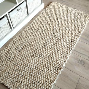 May include: A rectangular area rug featuring a chunky, hand-knit texture in an off-white, cream color. The rug is constructed from thick, braided yarn loops, creating a dense, uniform pattern across the entire surface of the floor covering.