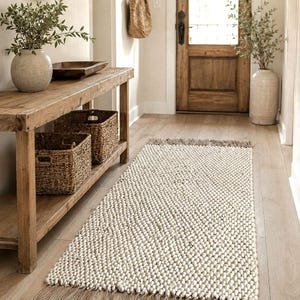 A rectangular runner rug featuring a thick, cream-colored chunky  texture with a prominent braided weave pattern. The rug is finished with long, thin, natural-toned jute fringe tassels along both of the shorter horizontal edges.