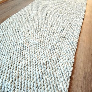 A long, rectangular runner rug featuring a thick, chunky knit texture in a light cream and off-white color palette. The rug is constructed with dense, rounded yarn loops arranged in a consistent, repeating pattern across the surface.