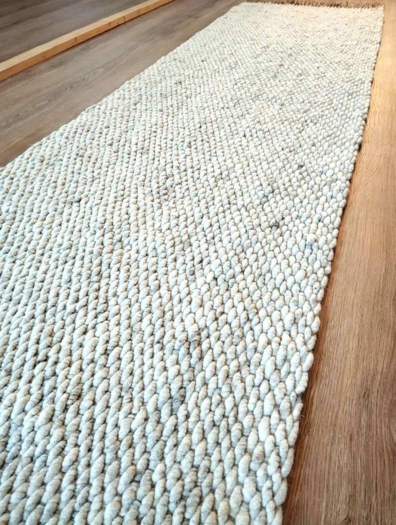 A long, rectangular runner rug featuring a thick, chunky knit texture in a light cream and off-white color palette. The rug is constructed with dense, rounded yarn loops arranged in a consistent, repeating pattern across the surface.