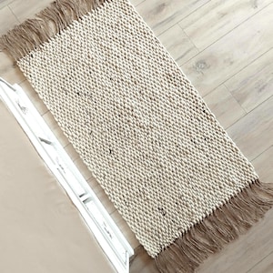May include: A rectangular area rug featuring a thick, cream-colored knit texture with subtle dark flecks throughout. The rug is finished with long, light brown fringe tassels on both shorter ends, creating a distinct contrast in color and texture.