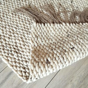 A wool jute runner rug featuring a thick, chunky texture in a light cream and off-white color. The rug is folded over itself, revealing the dense, looped yarn construction and a fringed edge.