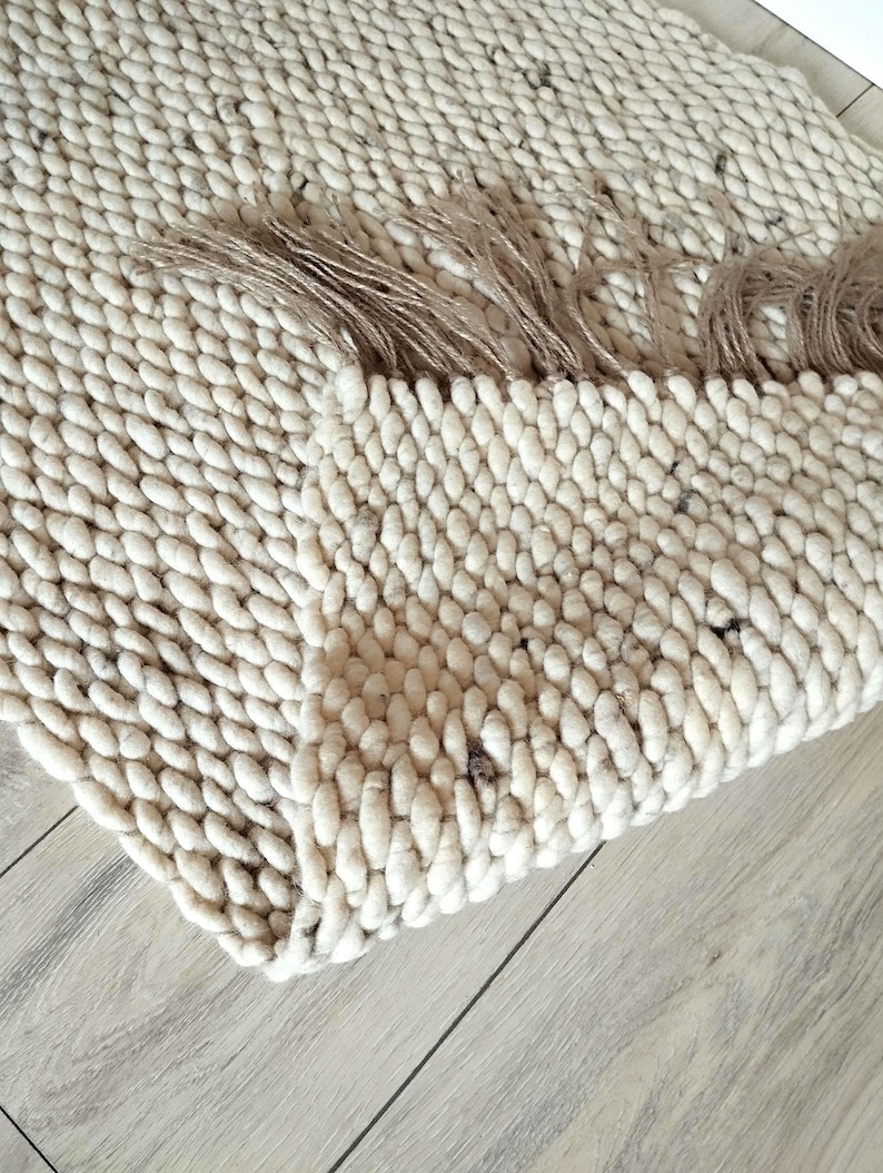 A wool jute runner rug featuring a thick, chunky texture in a light cream and off-white color. The rug is folded over itself, revealing the dense, looped yarn construction and a fringed edge.