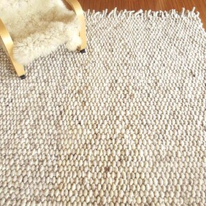 Natural undyed chunky wool rug, handwoven with thick textured loops and fringe, shown on a wooden floor in soft neutral tones.