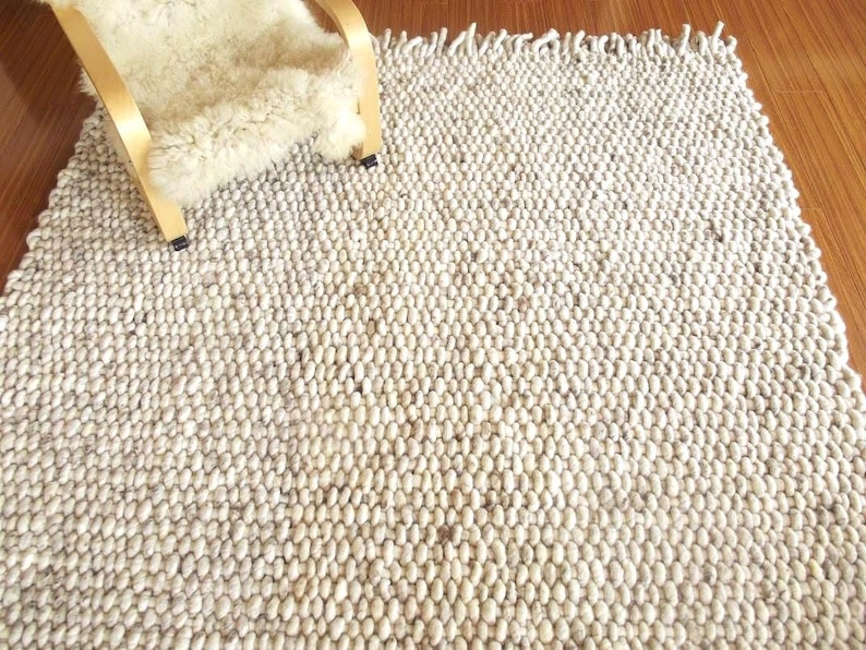 Natural undyed chunky wool rug, handwoven with thick textured loops and fringe, shown on a wooden floor in soft neutral tones.