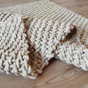 May include: A chunky knit blanket in a solid cream colour, featuring a thick, oversized yarn texture with a consistent interlocking stitch pattern throughout the entire piece, creating a soft, voluminous, and heavy appearance across the fabric.
