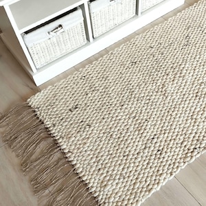 May include: A rectangular area rug featuring a thick, chunky knit weave in an off-white, cream-colored yarn with subtle dark flecks throughout. The rug is finished with long, natural-toned jute or twine fringe along the short edges.