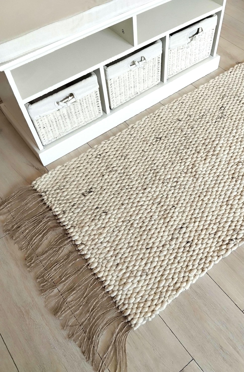 May include: A rectangular area rug featuring a thick, chunky knit weave in an off-white, cream-colored yarn with subtle dark flecks throughout. The rug is finished with long, natural-toned jute or twine fringe along the short edges.