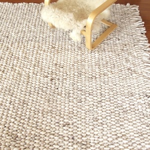 Handwoven chunky wool rug shown from an angle, with dense looped texture, fringe edge and natural cream/ ivory/ off-white and grey tones beside a lounge chair.
