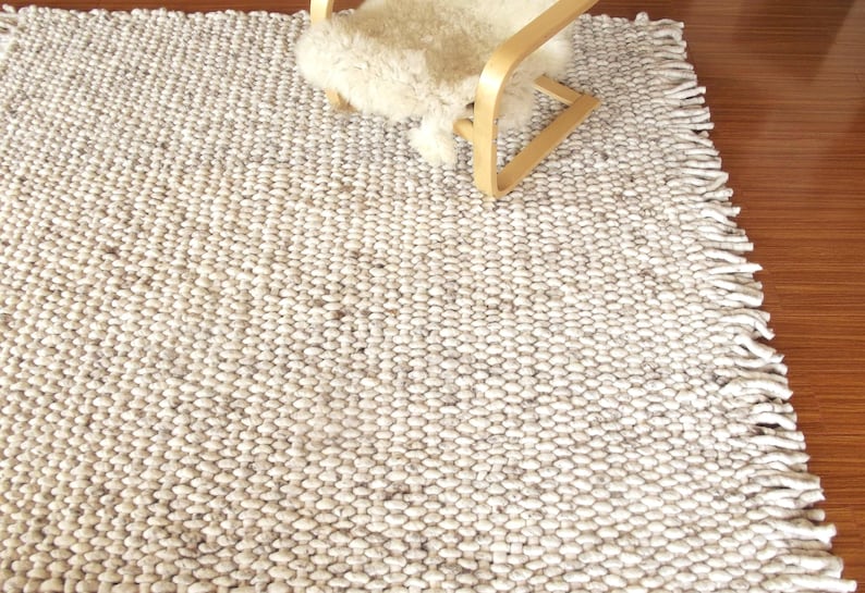 Handwoven chunky wool rug shown from an angle, with dense looped texture, fringe edge and natural cream/ ivory/ off-white and grey tones beside a lounge chair.