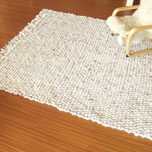 Wide view of a handwoven chunky wool area rug in natural off-white and grey tones, with thick looped texture and fringe, styled on a wooden floor beside a light lounge chair.