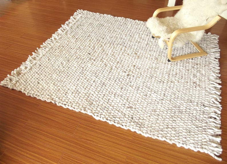 Wide view of a handwoven chunky wool area rug in natural off-white and grey tones, with thick looped texture and fringe, styled on a wooden floor beside a light lounge chair.