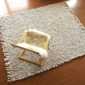 High-angle view of a handwoven chunky wool rug in natural ivory/ cream/ off-white and grey tones, with thick woven texture and fringe edges, styled on a wooden floor with a lounge chair for scale.