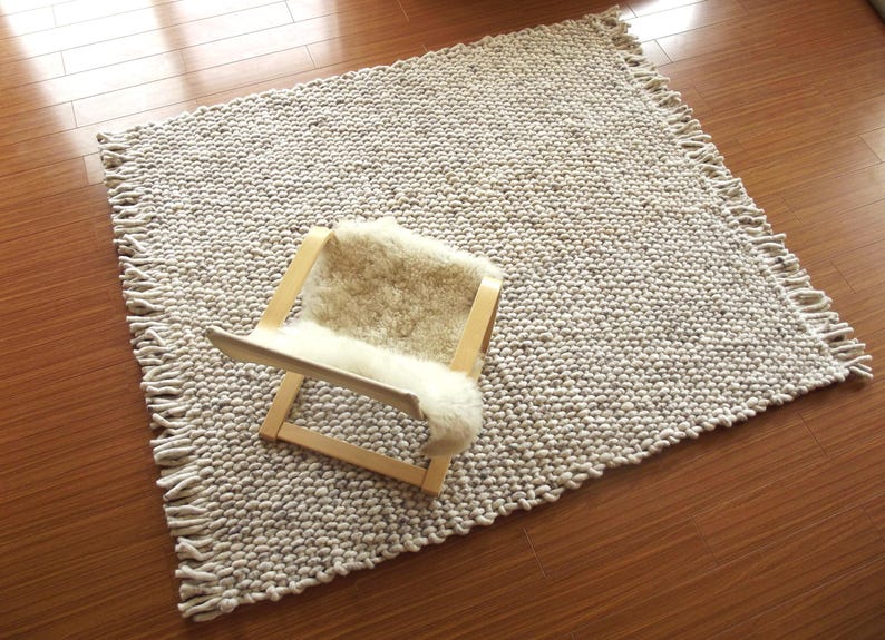 High-angle view of a handwoven chunky wool rug in natural ivory/ cream/ off-white and grey tones, with thick woven texture and fringe edges, styled on a wooden floor with a lounge chair for scale.