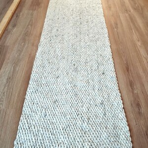 A long, rectangular runner rug featuring a thick, chunky texture in an off-white and light gray speckled color palette. The rug has a dense, braided weave pattern throughout its surface and is finished with a short, natural-toned fringe.