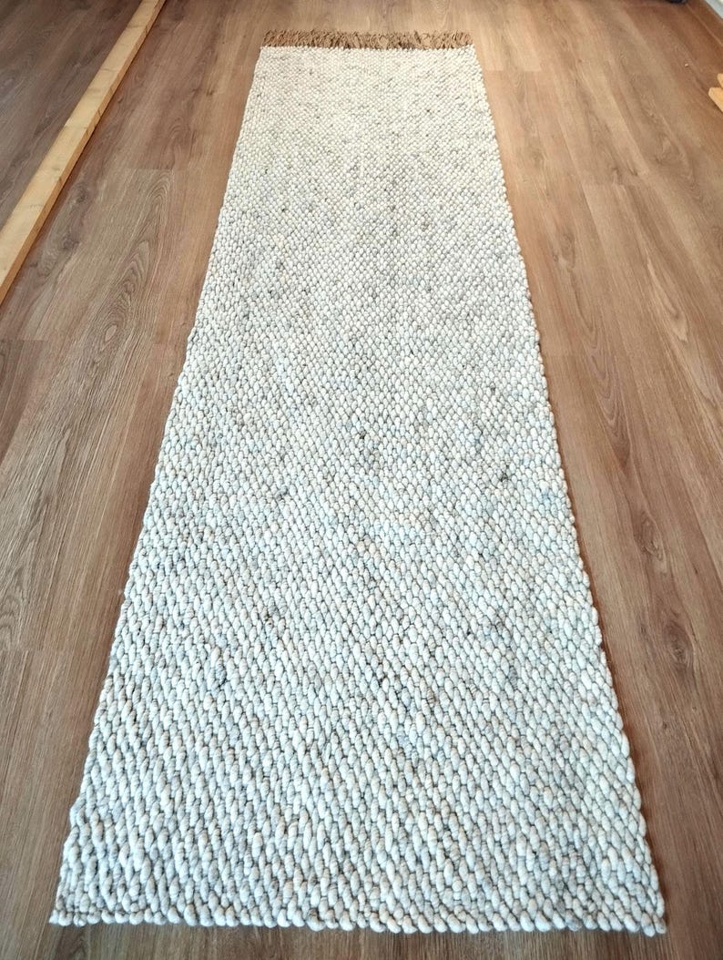 A long, rectangular runner rug featuring a thick, chunky texture in an off-white and light gray speckled color palette. The rug has a dense, braided weave pattern throughout its surface and is finished with a short, natural-toned fringe.