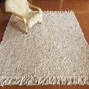Chunky wool rug, handwoven from natural undyed wool in cream/ off-white and soft grey tones, with thick texture and fringe, shown as a Scandinavian neutral area rug for living room or bedroom