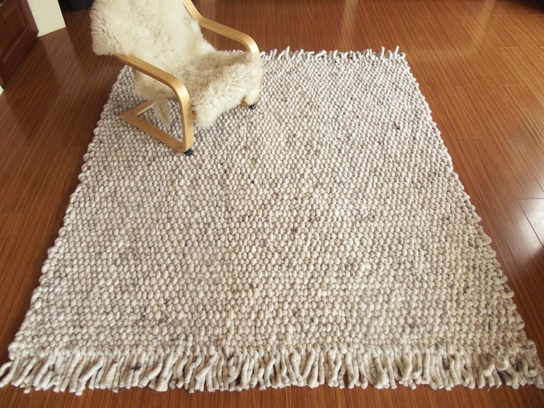 Chunky wool rug, handwoven from natural undyed wool in cream/ off-white and soft grey tones, with thick texture and fringe, shown as a Scandinavian neutral area rug for living room or bedroom