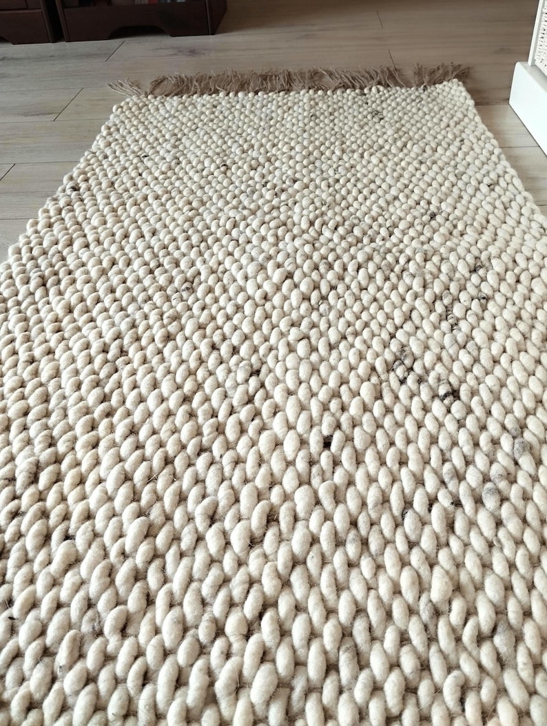 A long runner rug featuring a thick, chunky texture made from light beige/ off-white yarn with dark flecks. The rug displays a consistent, repeating braided pattern across its entire surface, creating a dense and tactile appearance.
