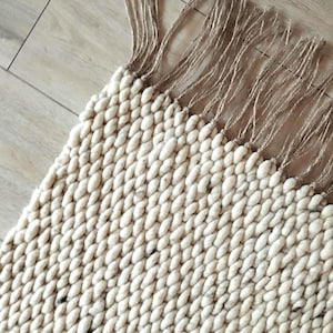Close up of a corner of a wool jute runner rug featuring a thick, chunky texture in a light cream and off-white color palette, with dark speckles. The surface is composed of densely woven, rounded yarn loops that create a uniform, tactile, and highly textured appearance.