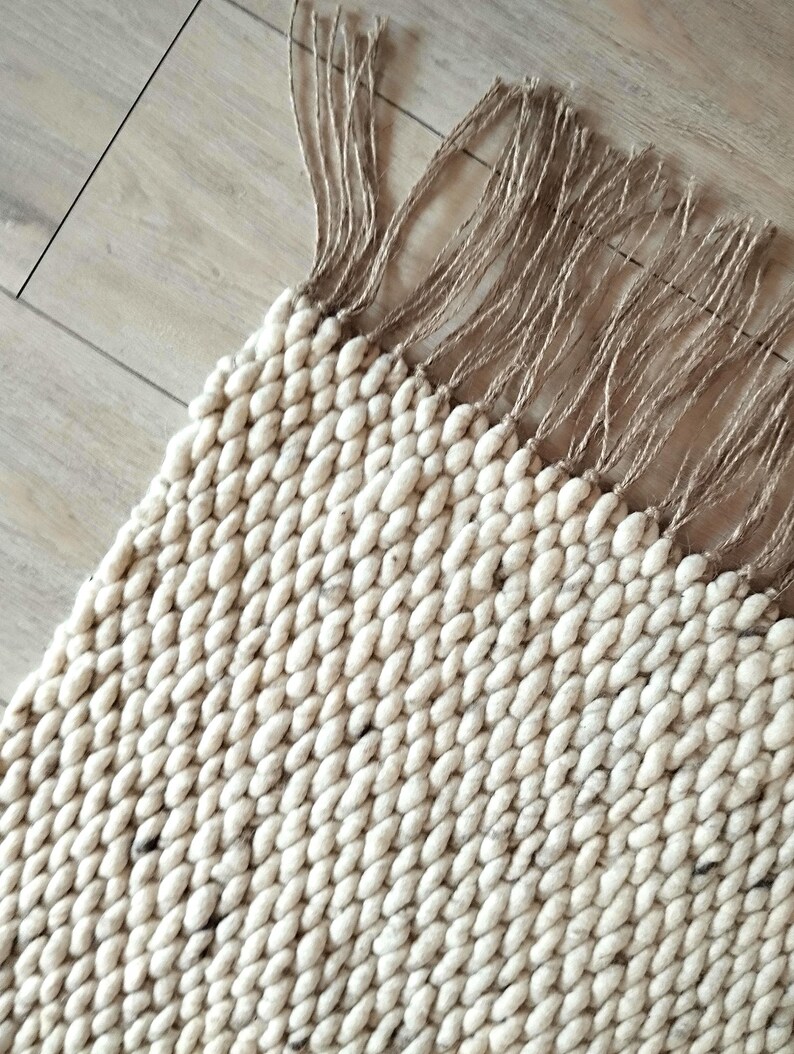 Close up of a corner of a wool jute runner rug featuring a thick, chunky texture in a light cream and off-white color palette, with dark speckles. The surface is composed of densely woven, rounded yarn loops that create a uniform, tactile, and highly textured appearance.