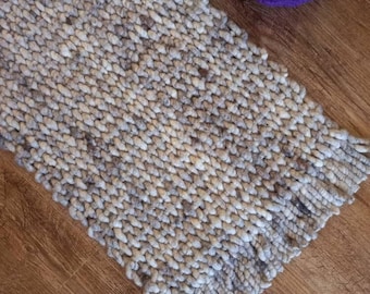 Peut inclure: Tapis fait à la main, texturé, dans des tons de gris et de beige, avec une bordure à franges. Deux pelotes de laine, une blanche et une violette, sont placées près du tapis. La texture épaisse et bouclée du tapis suggère un aspect artisanal et confortable.