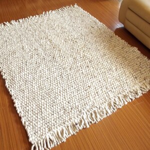 Handwoven chunky wool rug in natural undyed off-white and soft grey tones, shown on a wooden floor with thick texture and  fringe