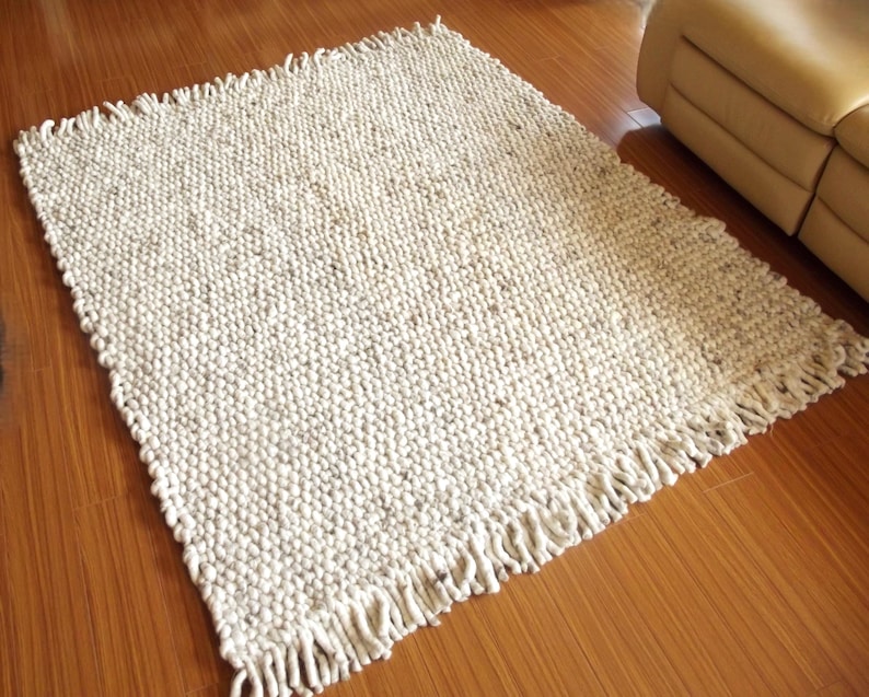 Handwoven chunky wool rug in natural undyed off-white and soft grey tones, shown on a wooden floor with thick texture and  fringe