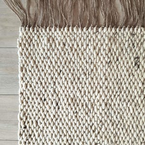 Close up of a corner of a woven runner rug featuring a textured off-white with dark speckles pattern. The top edge is finished with a row of natural tan jute-colored fringe.