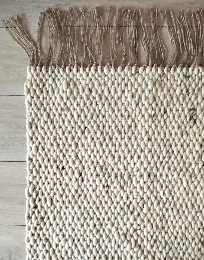 Close up of a corner of a woven runner rug featuring a textured off-white with dark speckles pattern. The top edge is finished with a row of natural tan jute-colored fringe.