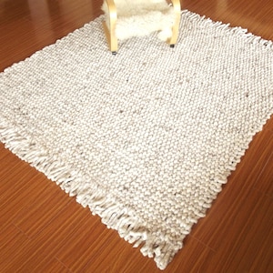 Full view of a handwoven chunky wool rug with thick woven texture and fringe edges, in natural cream/ ivory/ off-white and grey tones, shown on a wooden floor with a chair for scale.