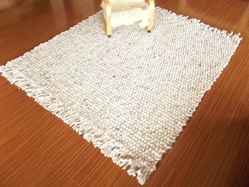 Full view of a handwoven chunky wool rug with thick woven texture and fringe edges, in natural cream/ ivory/ off-white and grey tones, shown on a wooden floor with a chair for scale.
