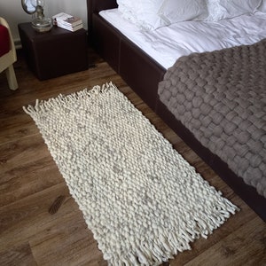 May include: A white and gray chunky knit rug with long fringe on a wooden floor. The rug is in front of a brown leather bed with white bedding and a brown knitted throw blanket.